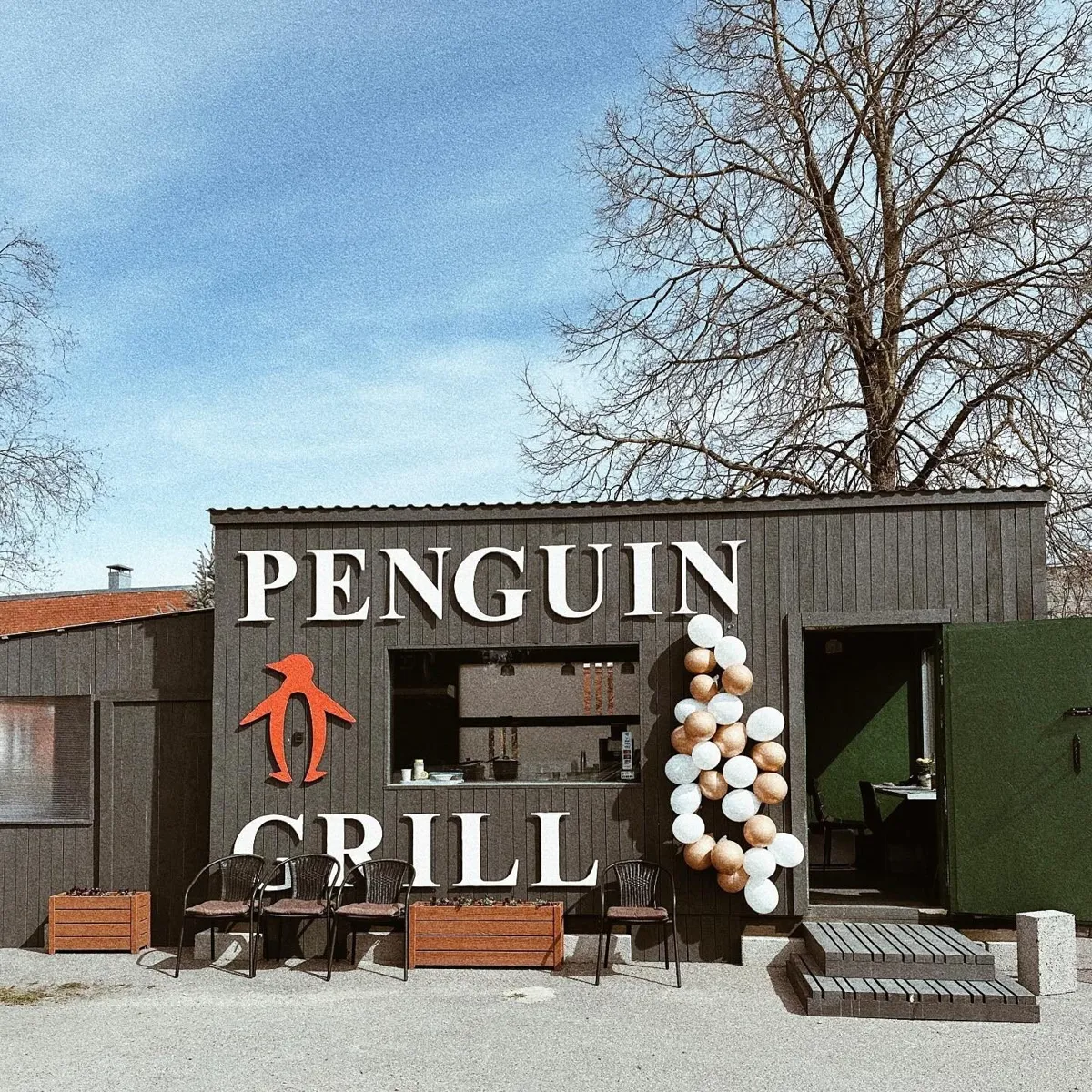 Penguin Grill kafejnīca&nbsp;– tumša koka ēka ar baltu uzrakstu, sarkanu pingvīnu un krēsliem ārā